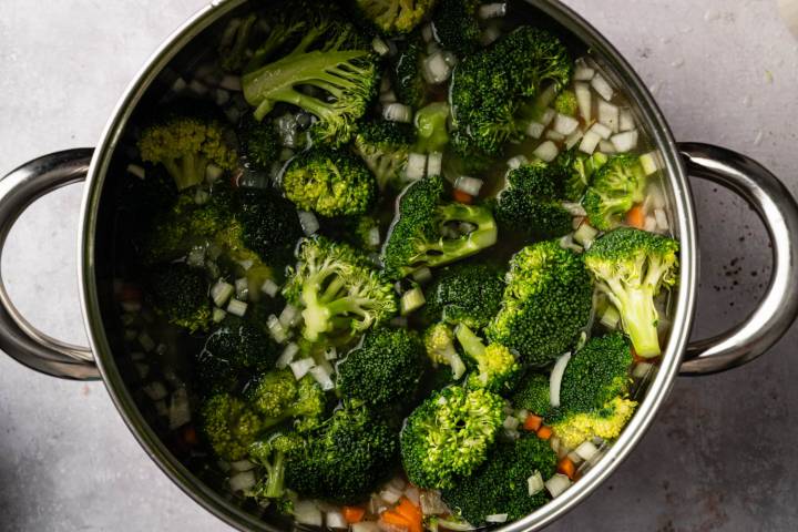 A pot filled with vibrant green broccoli florets, chopped onions, and diced carrots in water, ready for cooking. 
